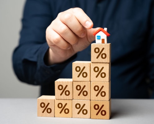 Houten blokken met procenttekens opgestapeld als een trap, met een miniatuurhuisje bovenop, als symbool voor stijgende hypotheekrente